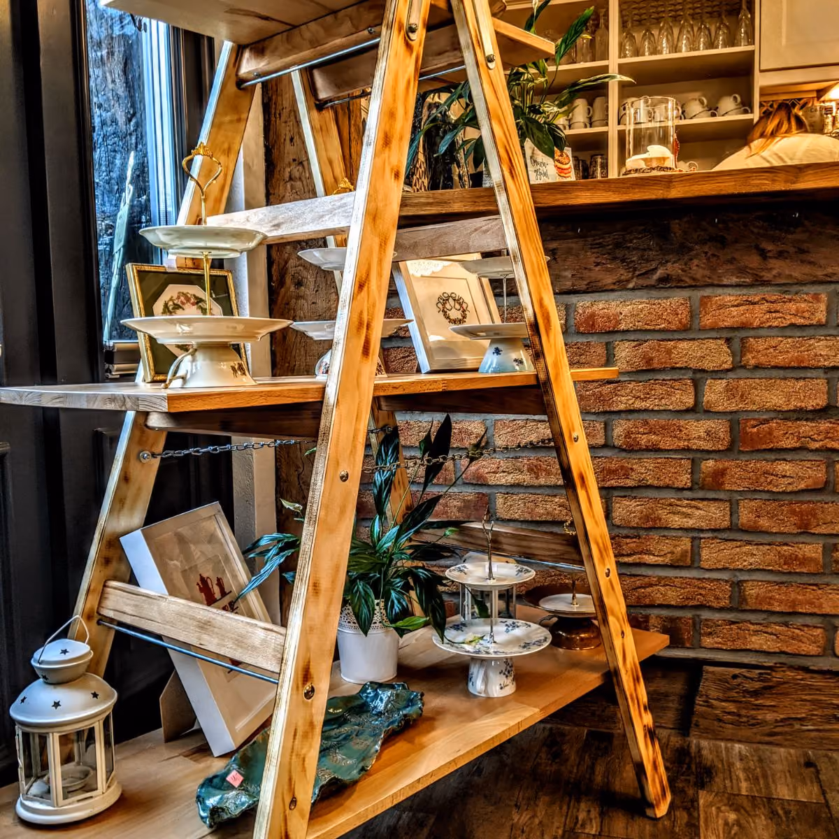 Wooden A-frame shelf with ceramic plates, framed pictures, plant, and lantern against brick wall.