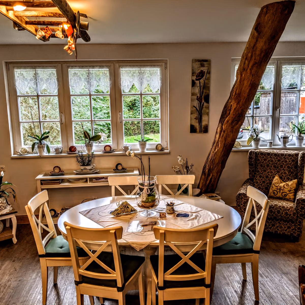 Cozy dining room with round table, six wooden chairs, large windows, and rustic wooden beam.