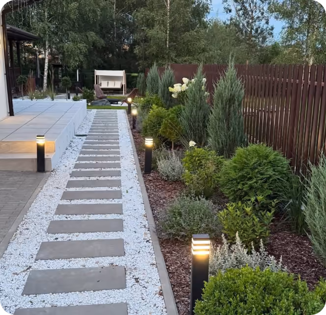 Stone slab path with white gravel and garden lights along green shrubs and trees by a wooden fence.