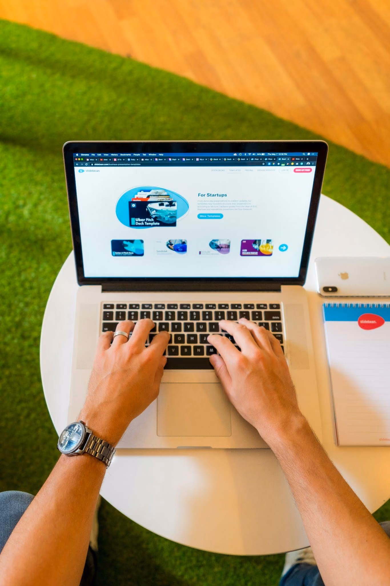 Person tippt auf einer Laptop-Tastatur, der Bildschirm zeigt eine Webseite mit Start-up Pitch-Deck-Vorlagen; auf dem weißen Tisch liegen ein iPhone und ein Notizblock.