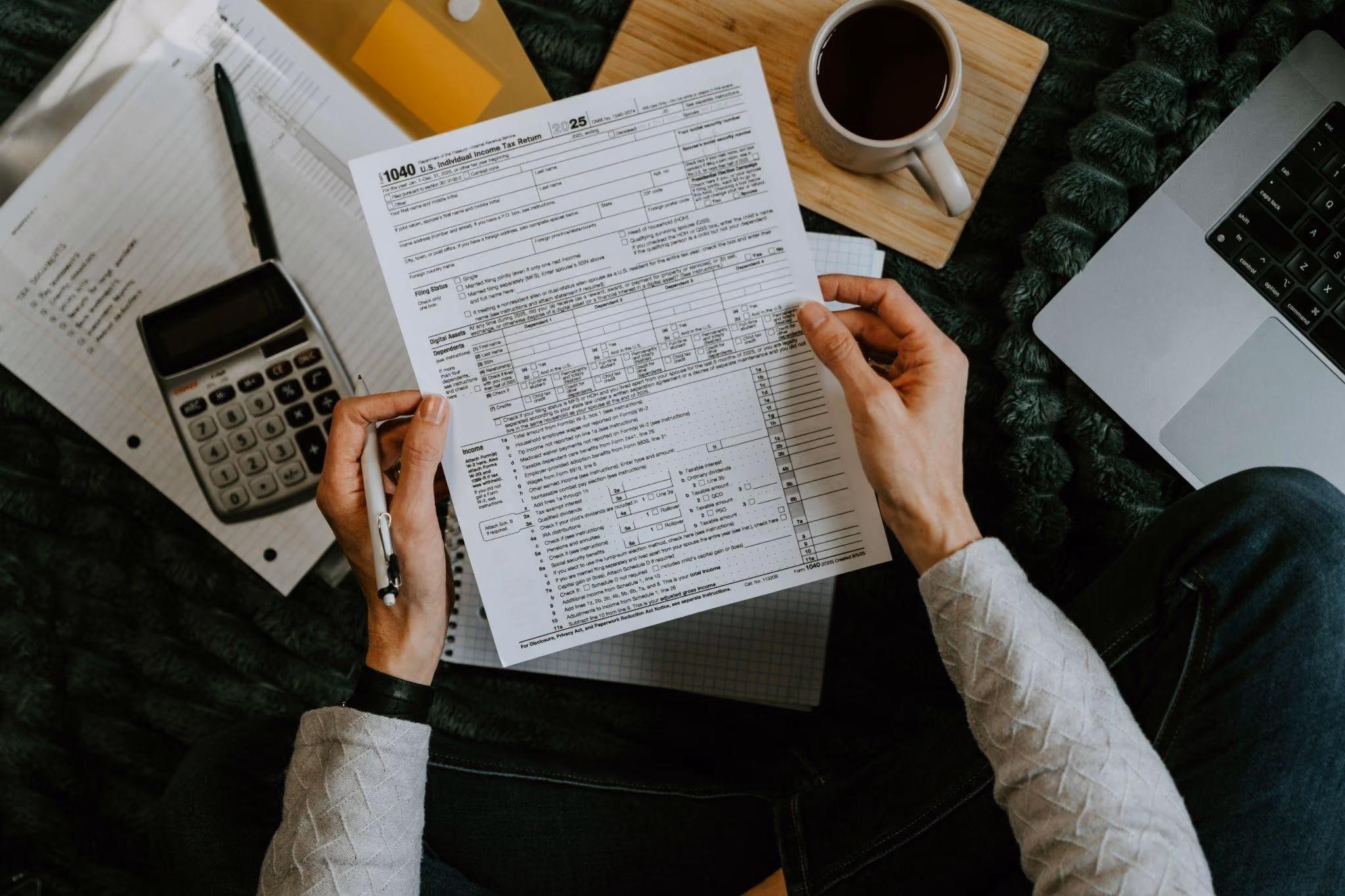 Person hält Formular 1040 zur US-Einkommensteuererklärung 2025 mit einem Stift in der Hand, umgeben von Taschenrechner, Stift, Kaffeetasse auf Holzunterlage und Laptop.