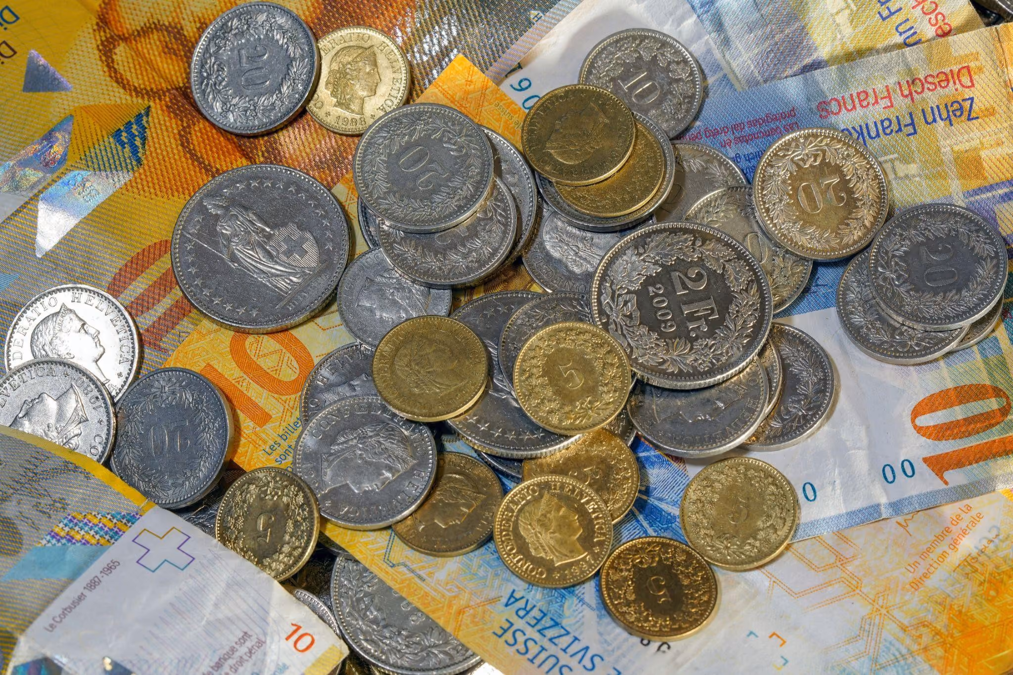 Verschiedene Schweizer Münzen aus Silber und Gold, darunter 2-Franken- und 20-Rappen-Münzen, verteilt auf mehreren bunten Schweizer Banknoten mit Werten von 10 und 100 Franken.