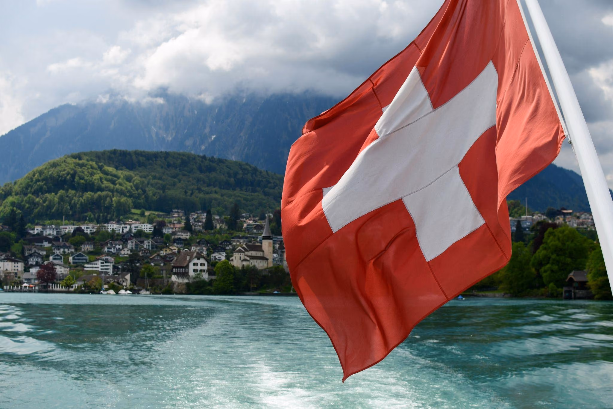 Schweizer Flagge weht an einem Boot mit Blick auf einen See, eine Stadt am Ufer mit Häusern und einer Kirche, bewaldete Hügel und Berge unter bewölktem Himmel.
