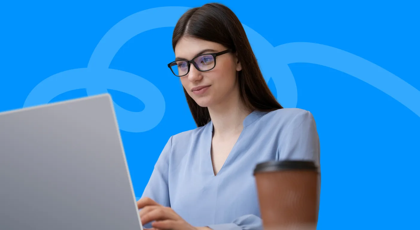 Woman in glasses working at a laptop next to a takeaway coffee cup.