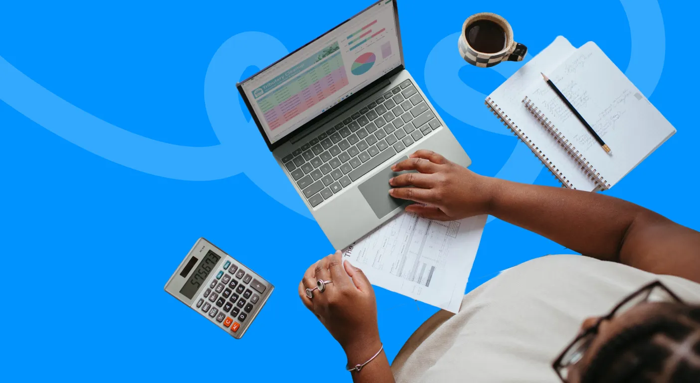 Overhead view of a person working at a laptop showing sales charts, with a calculator, notepad and coffee on the desk.