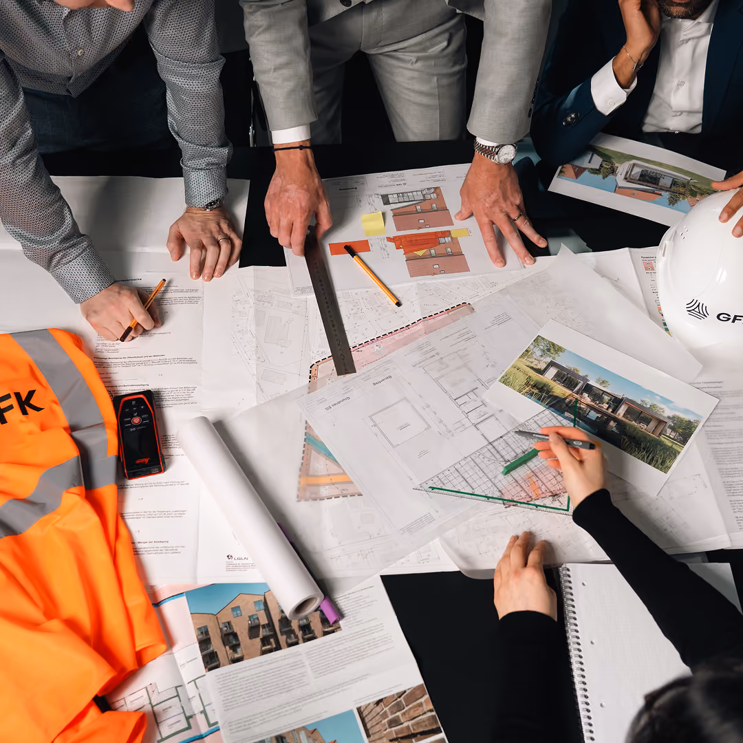 Architekten arbeiten an Bauplänen mit Lineal, Stiften, Bauhelm und orangener Sicherheitsweste auf Tisch.