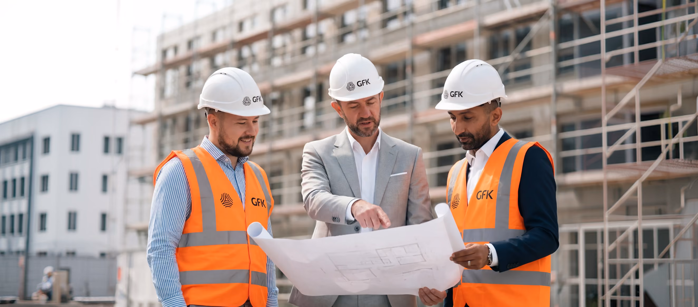 Drei Männer mit GFK-Helmen und Warnwesten besprechen Pläne vor einer Baustelle.