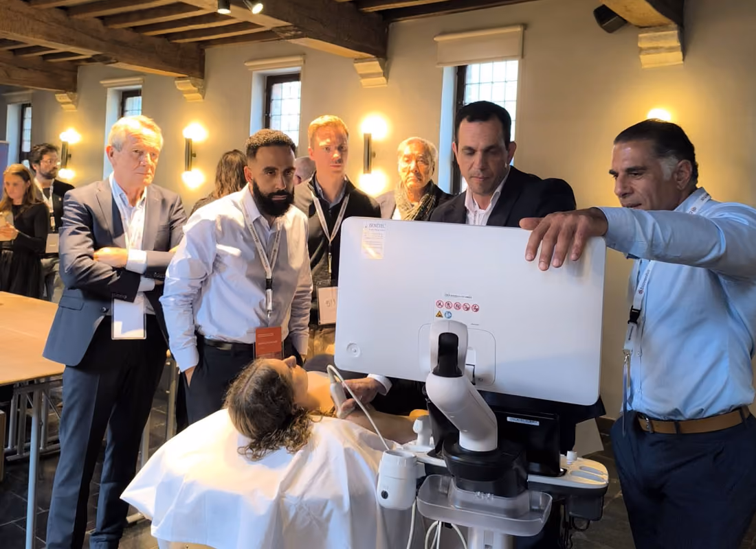 A group of professionals observes a medical ultrasound scan on a monitor with a patient lying on a bed.
