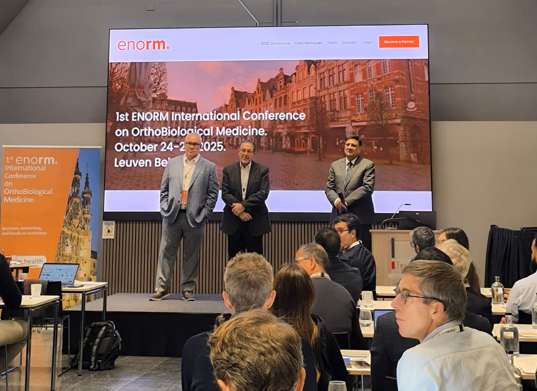Three men stand on stage in front of a crowd at the 1st ENORM International Conference on OrthoBiological Medicine.