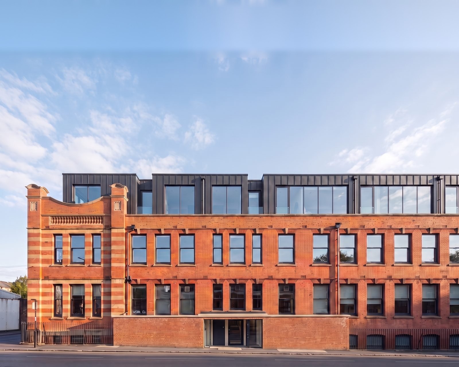 Enhanced photograph of the converted 1906 works at Foleshill Road, Coventry: the retained red-brick and terracotta facade below a new dark zinc-clad top storey.