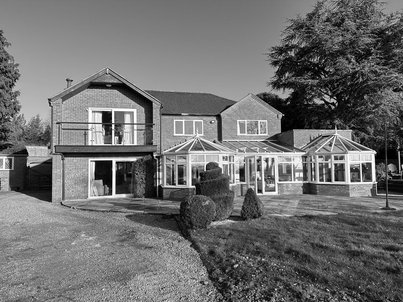 Black and white photograph of the existing house before the works: a two-storey brick dwelling with a first-floor balcony and two conservatories across the rear elevation, with lawn and mature trees around it