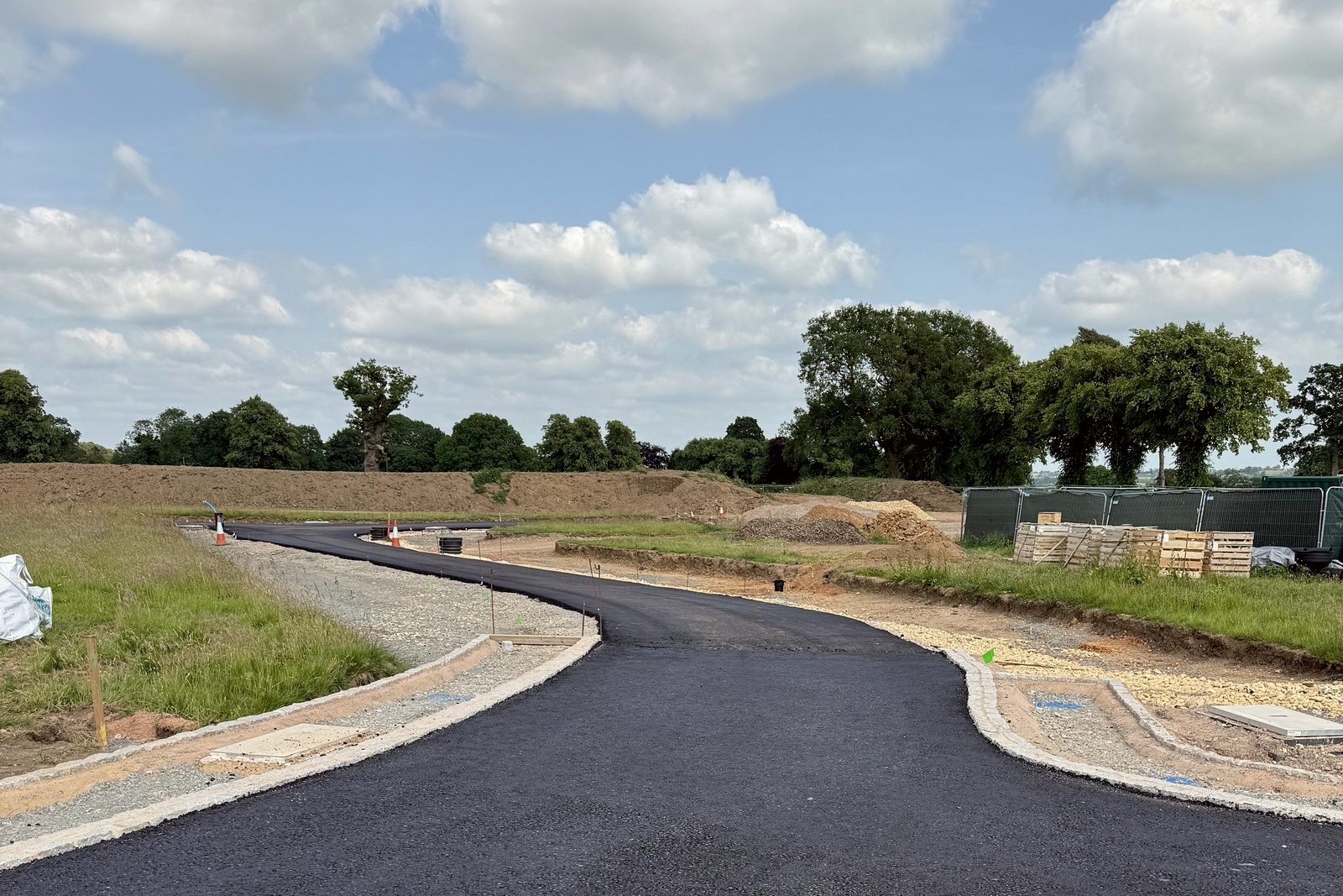 Photograph of the new estate road at Inwoods Park, Rugby, June 2025: fresh tarmac curving between kerbed plot entrances and grass verges, with site fencing, stacked materials and mature trees beyond