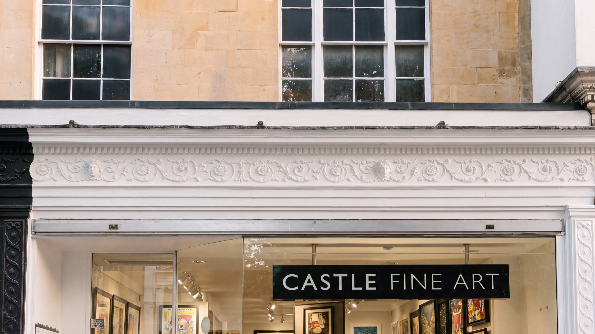 The Castle Fine Art gallery at 82A The Promenade, Cheltenham: the shopfront window full of framed art beneath the fascia, set under the rare ornate plaster frieze and the honey ashlar sash windows of the Grade II listed Regency building