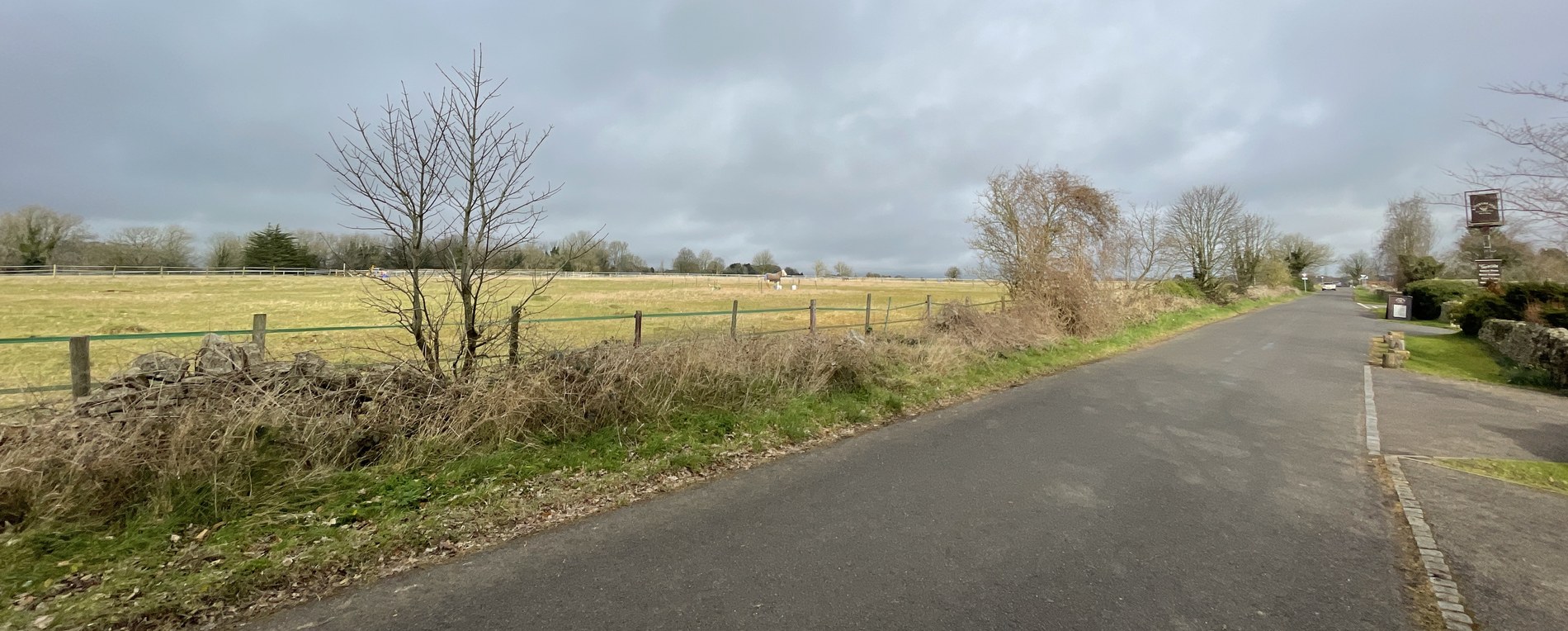 The approach to the site at Oakridge Lynch: a narrow lane running between a dry-stone wall and hedgerow on one side and a grass verge on the other, with a post-and-rail paddock, bare winter trees and open Cotswold fields stretching to a distant tree belt under a grey February sky