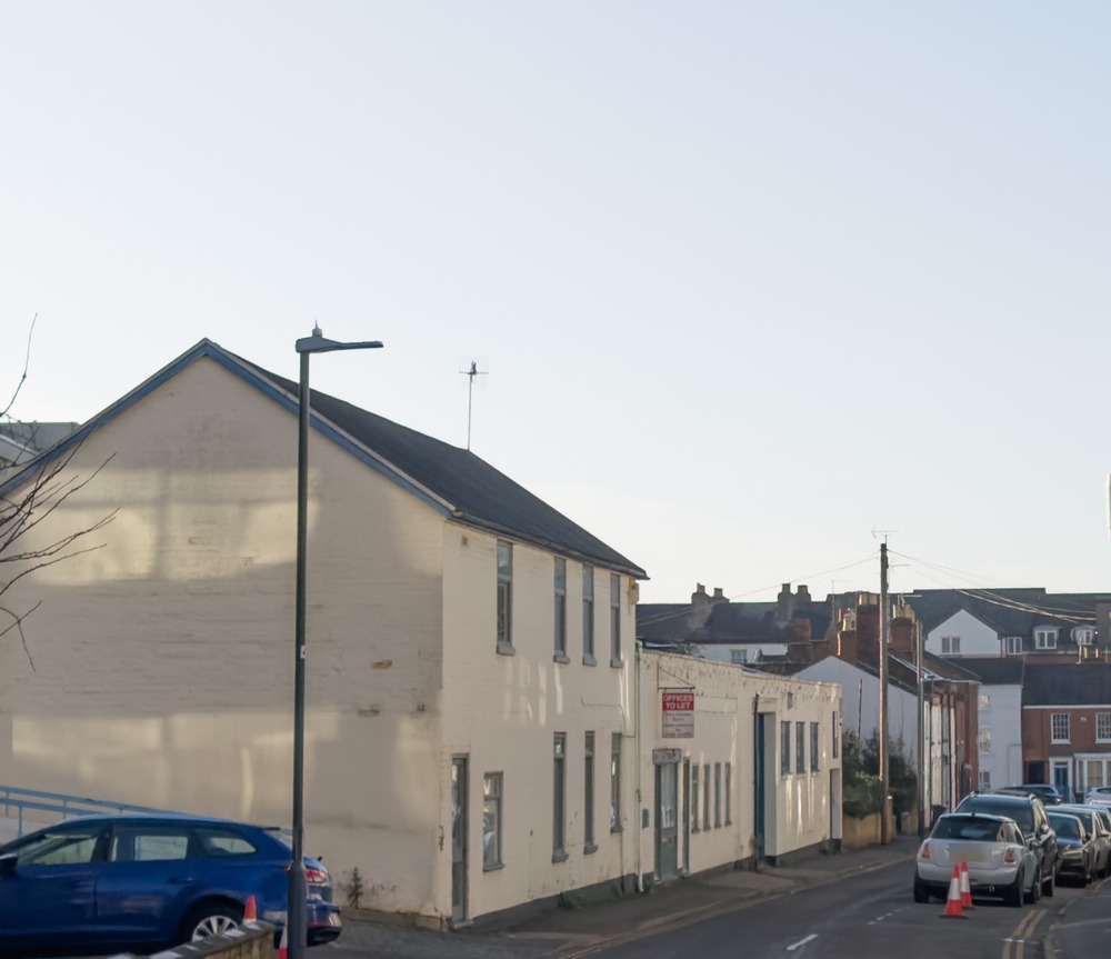The former commercial building at 16 Cross Street, Royal Leamington Spa before demolition, seen from the Willes Road junction: a cream painted two-storey building with a gable end, an offices to let sign, small windows and a loading door, with the narrow street and its terraced houses receding behind it
