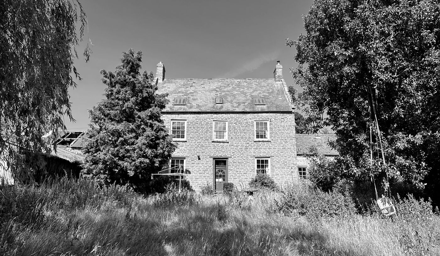 Black and white photograph of Barby Wood Farmhouse as found in 2023: the three-bay coursed ironstone farmhouse with twin brick stacks, twelve-pane sash windows in painted surrounds and a panelled front door, standing behind waist-high meadow grass with a willow to one side and a corrugated farm building to the other