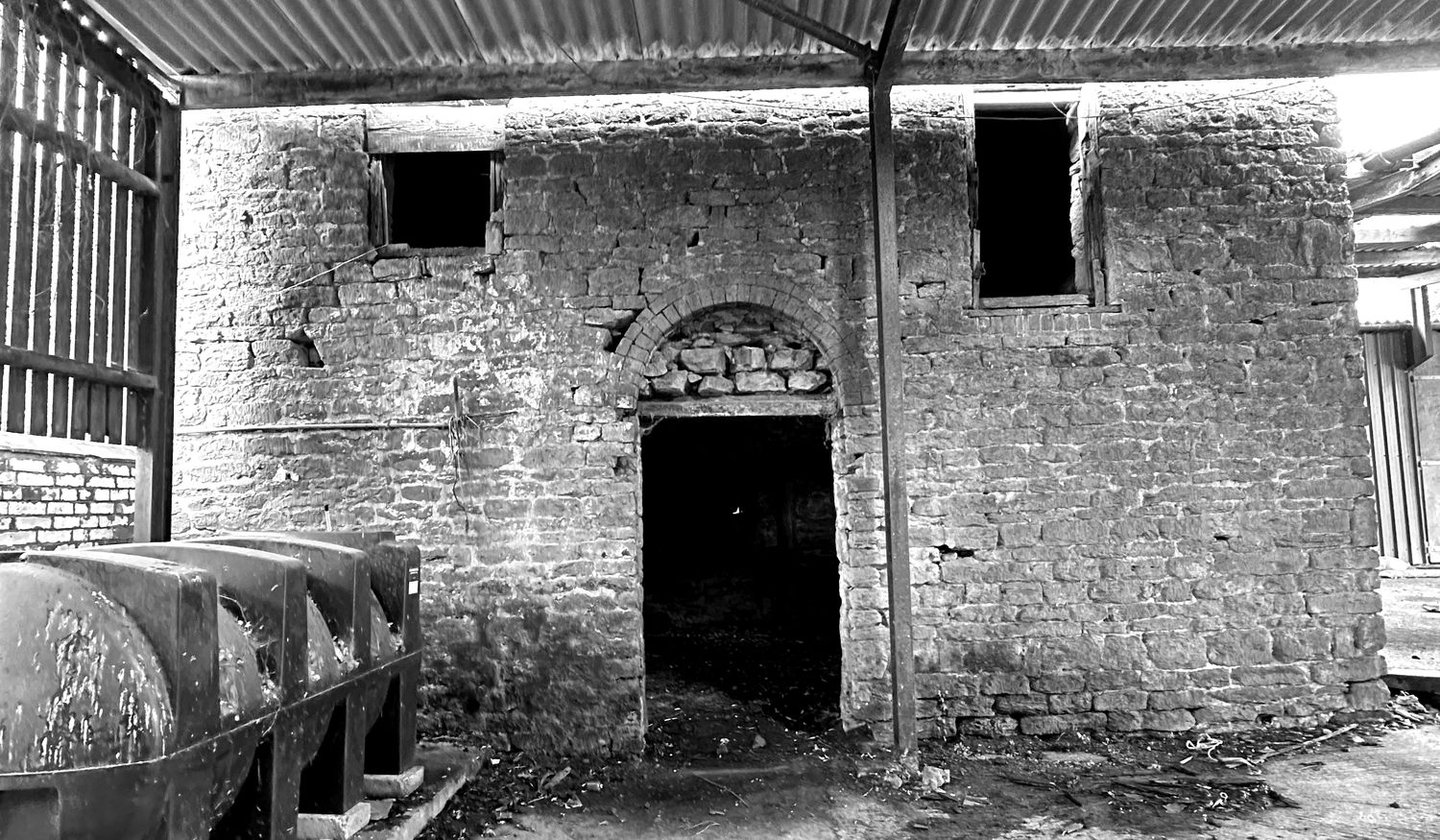 Black and white photograph of the grain barn at Barby Wood Farmhouse as found in 2023: a coursed ironstone elevation with a central doorway under a segmental brick arch and two small first-floor openings, seen from inside a later corrugated iron lean-to with plastic water tanks standing against the wall