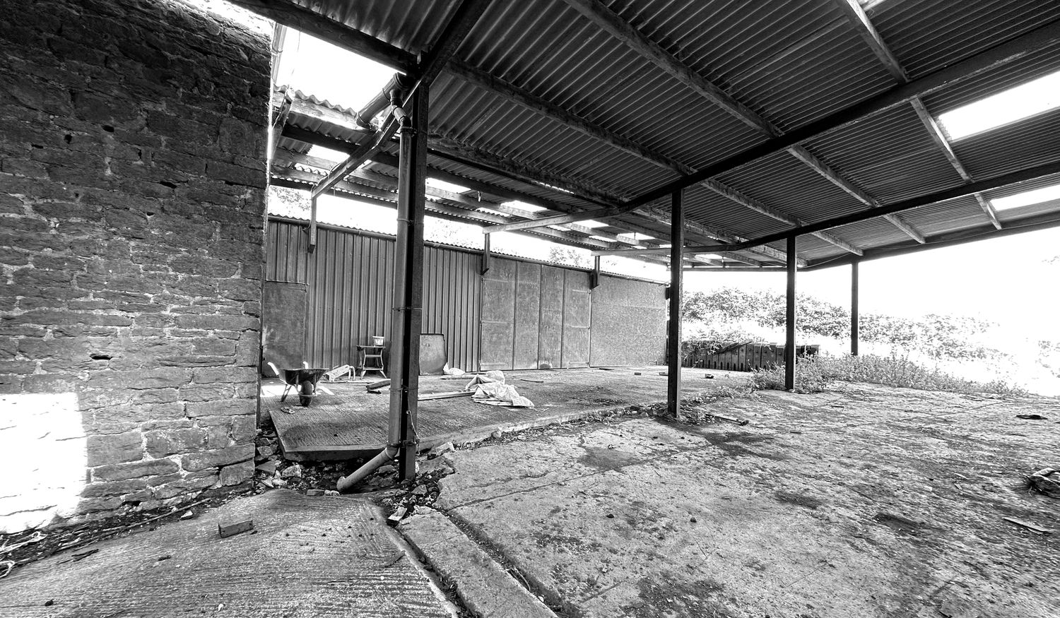Black and white photograph of later additions at Barby Wood Farmhouse as found in 2023: the gable of a coursed ironstone outbuilding with a first-floor opening, its ground floor closed off by a rusted corrugated sliding door and a breeze-block lean-to built against it, weeds growing through the concrete yard in front