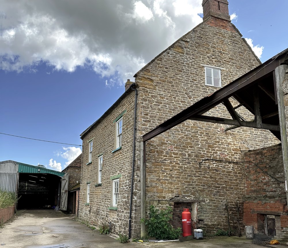 The gable end of the listed farmhouse at Barby Wood Farmhouse in 2023: coursed ironstone with a brick chimney stack and sash windows, with a modern steel-framed lean-to structure built hard against it and a concrete yard running away to a corrugated shed beyond