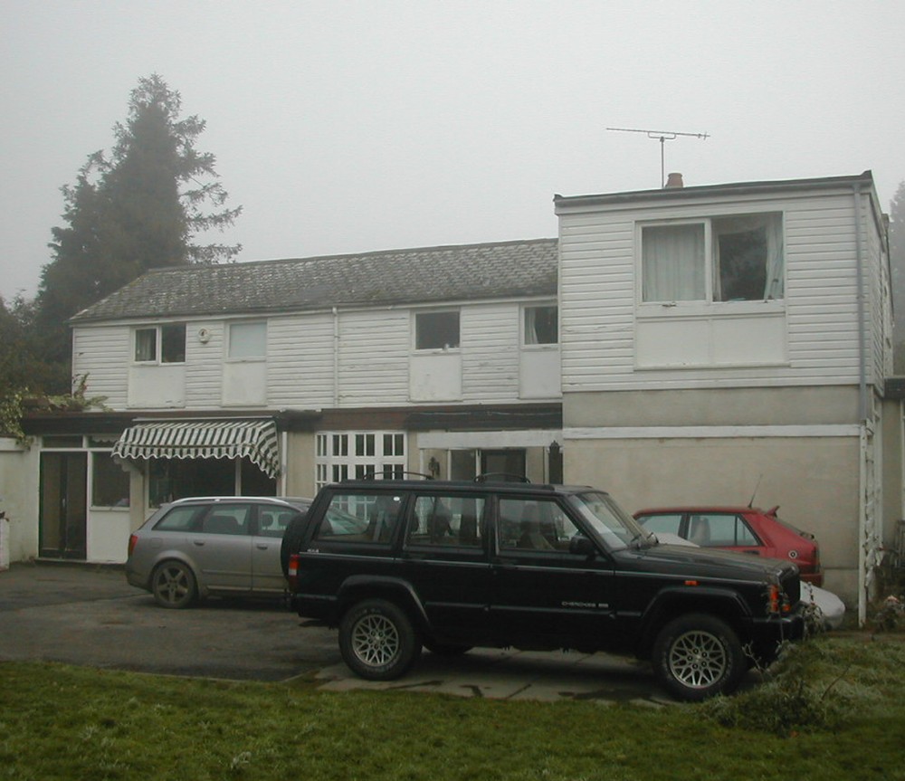 The post-war house that stood on the plot at Kenilworth Road, Royal Leamington Spa, photographed on a foggy morning before demolition: a tired two storey dwelling clad in white painted weatherboarding with a mossy tiled roof, a striped awning over the side door and cars parked on the hardstanding in front
