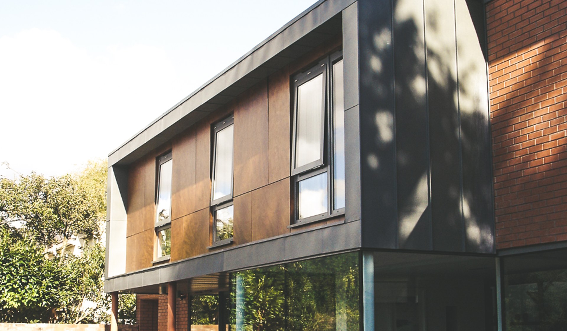The corner of the completed house on Kenilworth Road, Royal Leamington Spa: an upper storey of figured timber rainscreen panels and red facing brick wrapped in preweathered zinc, cantilevered over a fully glazed corner at ground level, with the dappled shadow of a mature tree across the zinc