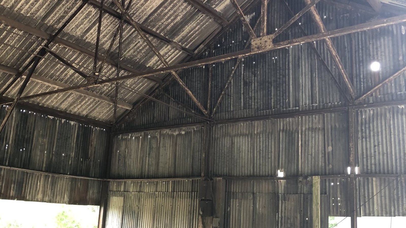 The inside of the redundant barn before conversion: a bolted steel truss roof over corrugated metal sheeting, daylight showing through gaps in the cladding, and farm machinery and clutter standing on the concrete floor.