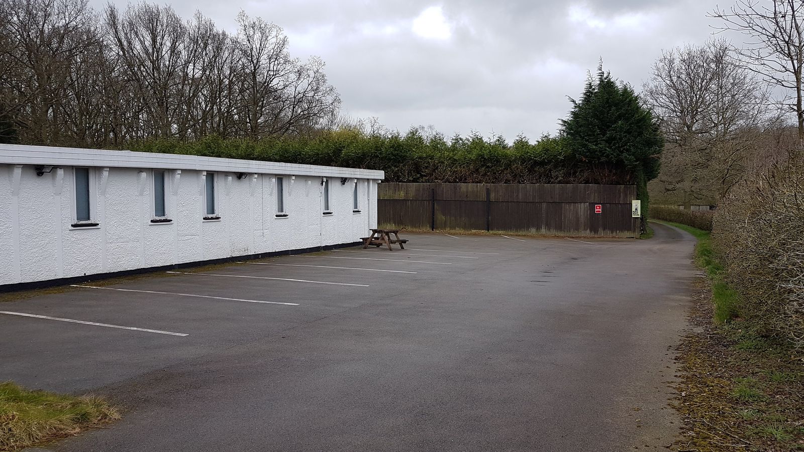 The site as found in March 2019: a long single-storey office building with a flat roof and painted white walls, standing behind a marked-out tarmac car park, with a close-boarded fence, clipped hedges and bare winter trees behind it.