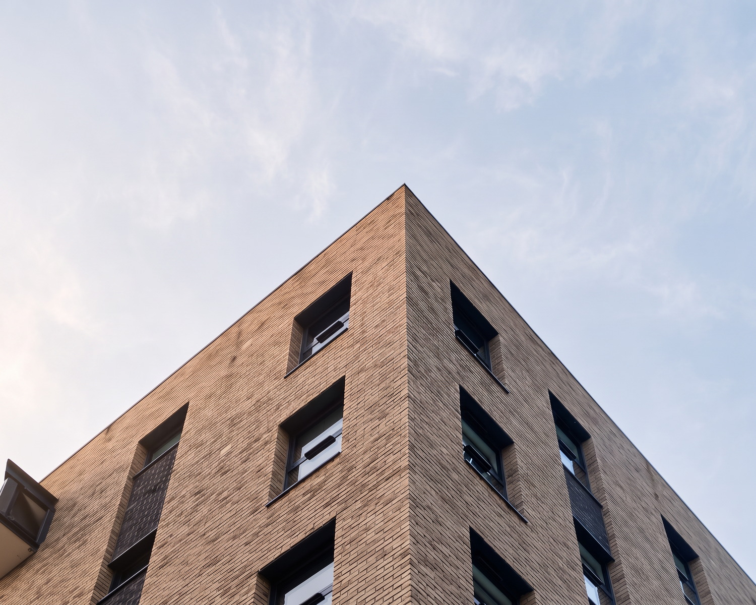 Enhanced photograph of the completed Tennant Street Lofts, Birmingham, looking up at the buff brick corner of the building in early morning light, its recessed windows stepping away on both elevations against a soft sky.