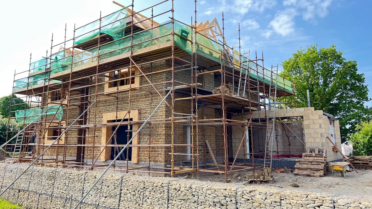 The replacement house nearing completion: a two-storey building in coursed golden-brown ironstone with gables, standing under scaffolding, with a stone-filled gabion retaining wall running across the foreground and a pallet of materials to one side.