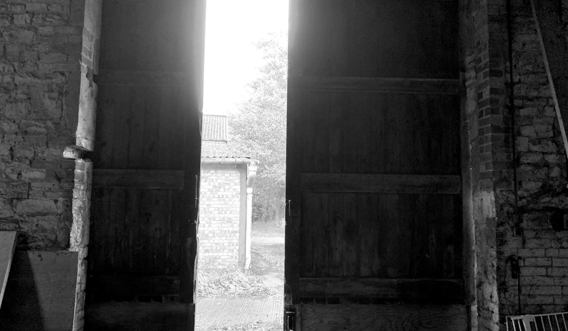 Black and white photograph from inside the barn as found, looking out through the open northern barn doors to the farmyard beyond