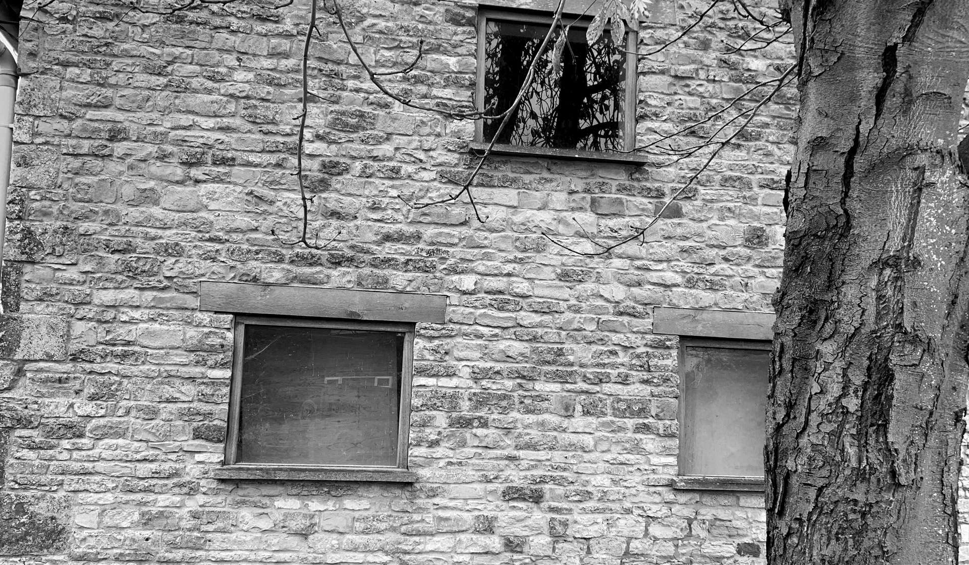 Black and white photograph of the barn's stone gable as found, with its original window openings boarded over and a mature tree in front of the coursed white lias wall