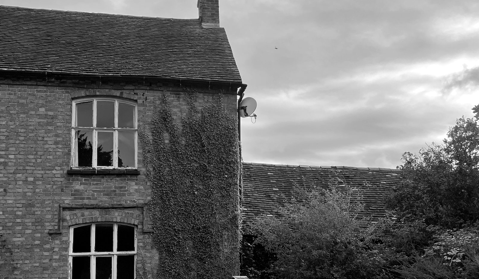 Black and white survey photograph of the farmhouse as found in 2021: a two-storey brick gable with multi-pane windows whose paint has failed, ivy growing across the wall, and the tiled roof of the attached farm building running away behind