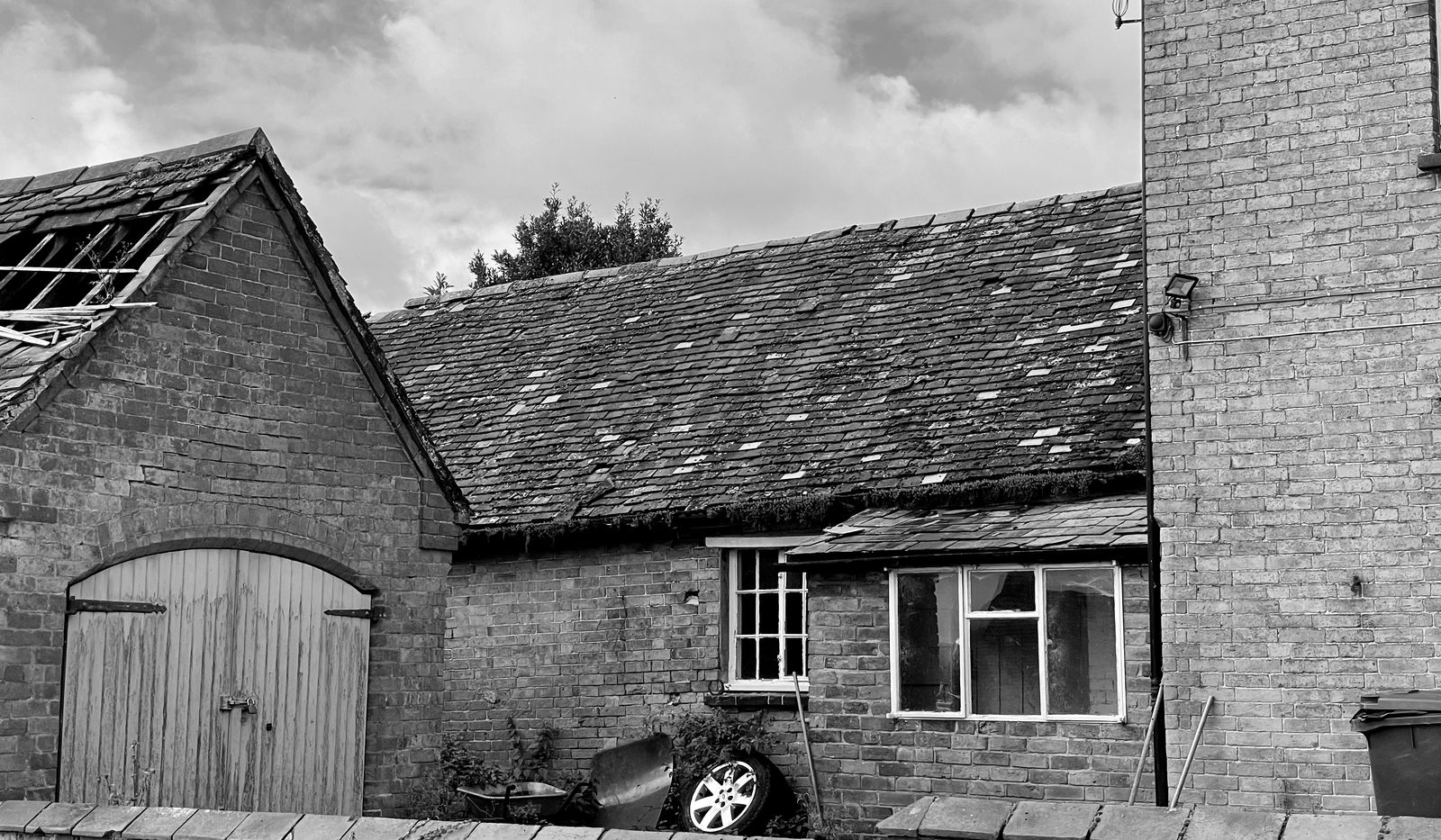Black and white survey photograph of the redundant farm building as found in 2021: a brick range with wide arched timber cart doors, a slipping and partly collapsed tiled roof at the near gable, running along to meet the brick gable of the farmhouse at the right