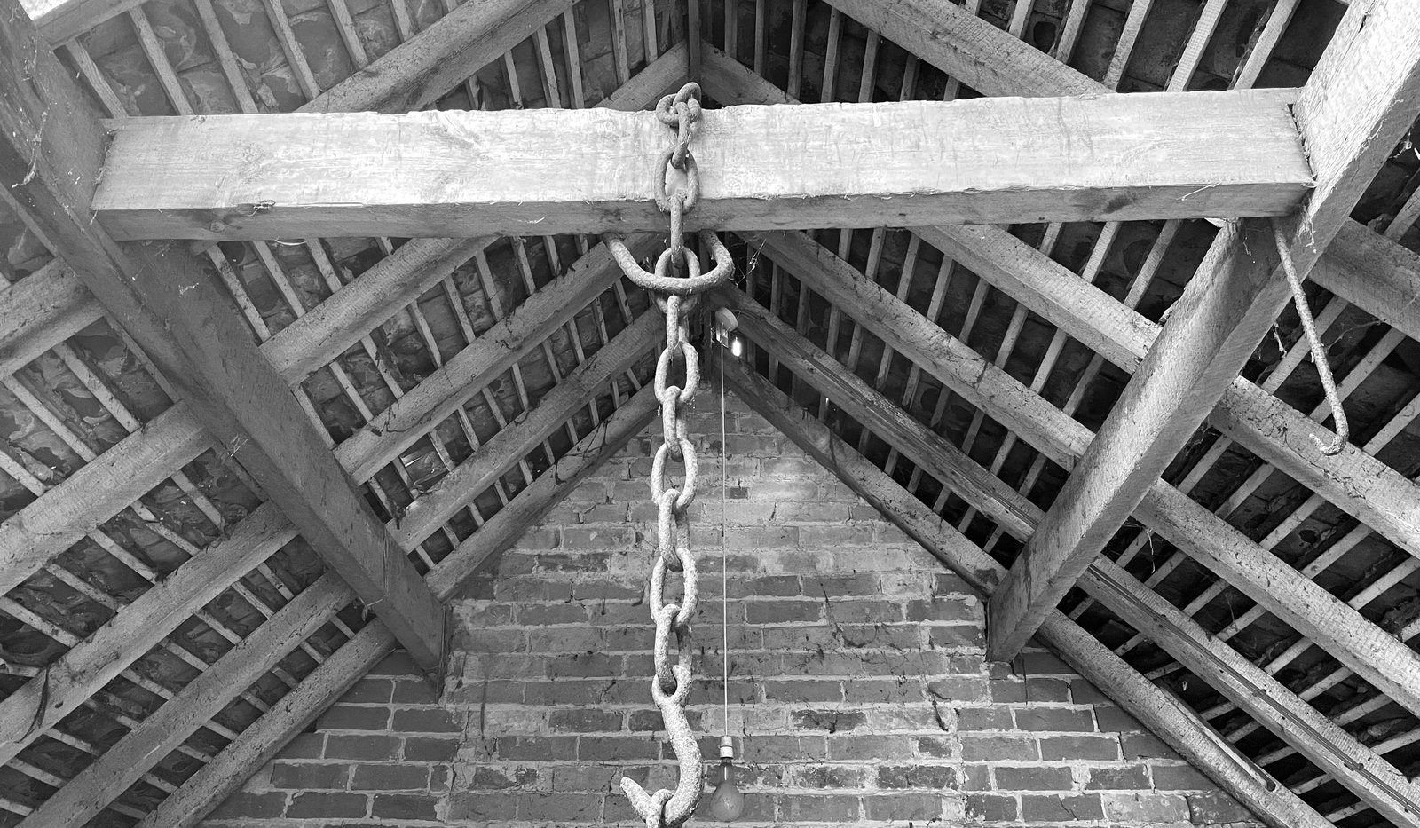 Black and white survey photograph looking up inside the farm building as found: a heavy timber tie beam and king post truss with rafters and battens above, a hoist chain hanging from the beam, and a brick gable wall behind