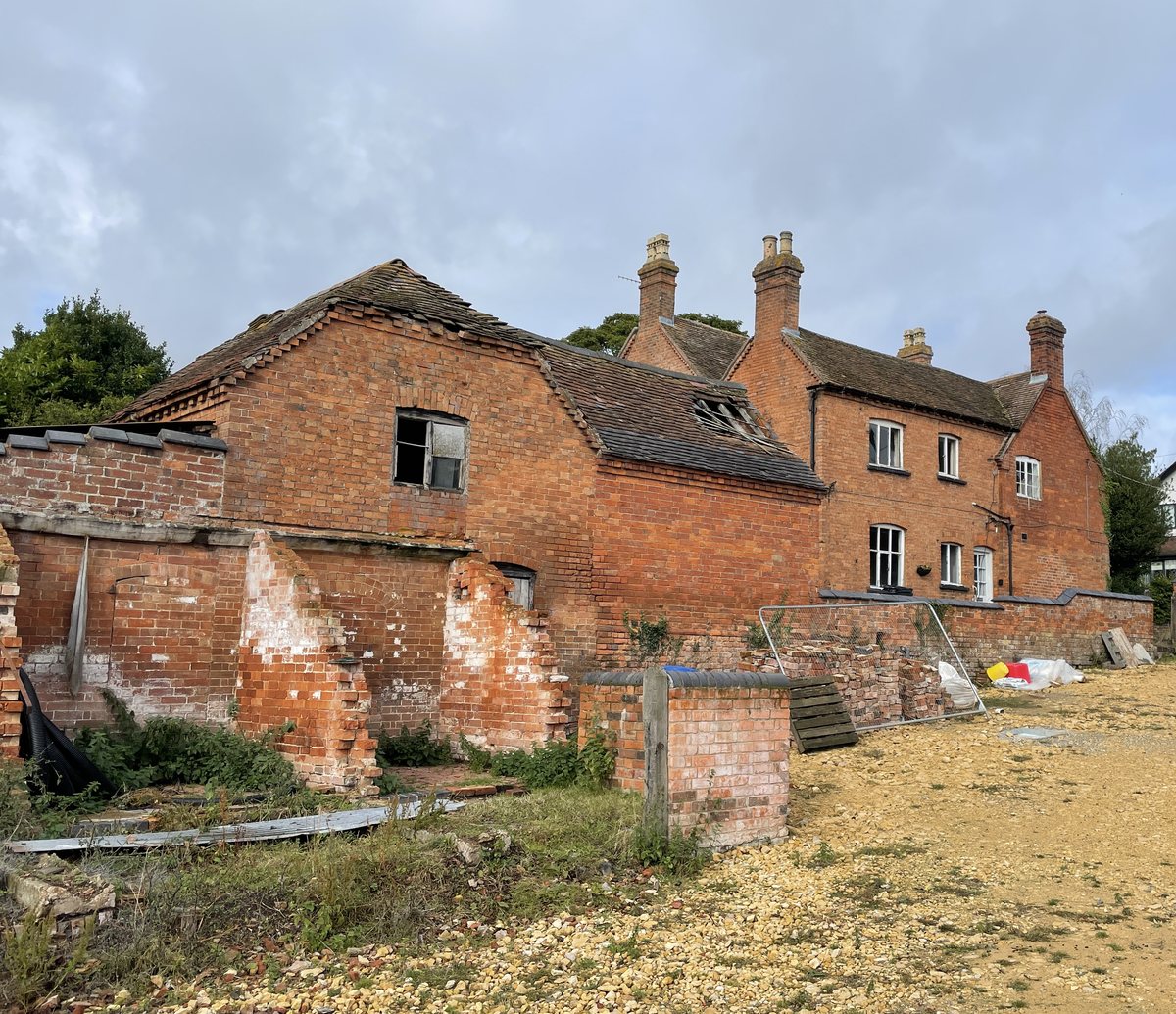 The farmyard as found, in colour: a ruined brick range in the foreground with its render falling away, the two-storey brick farmhouse standing behind it, and the village roofs beyond