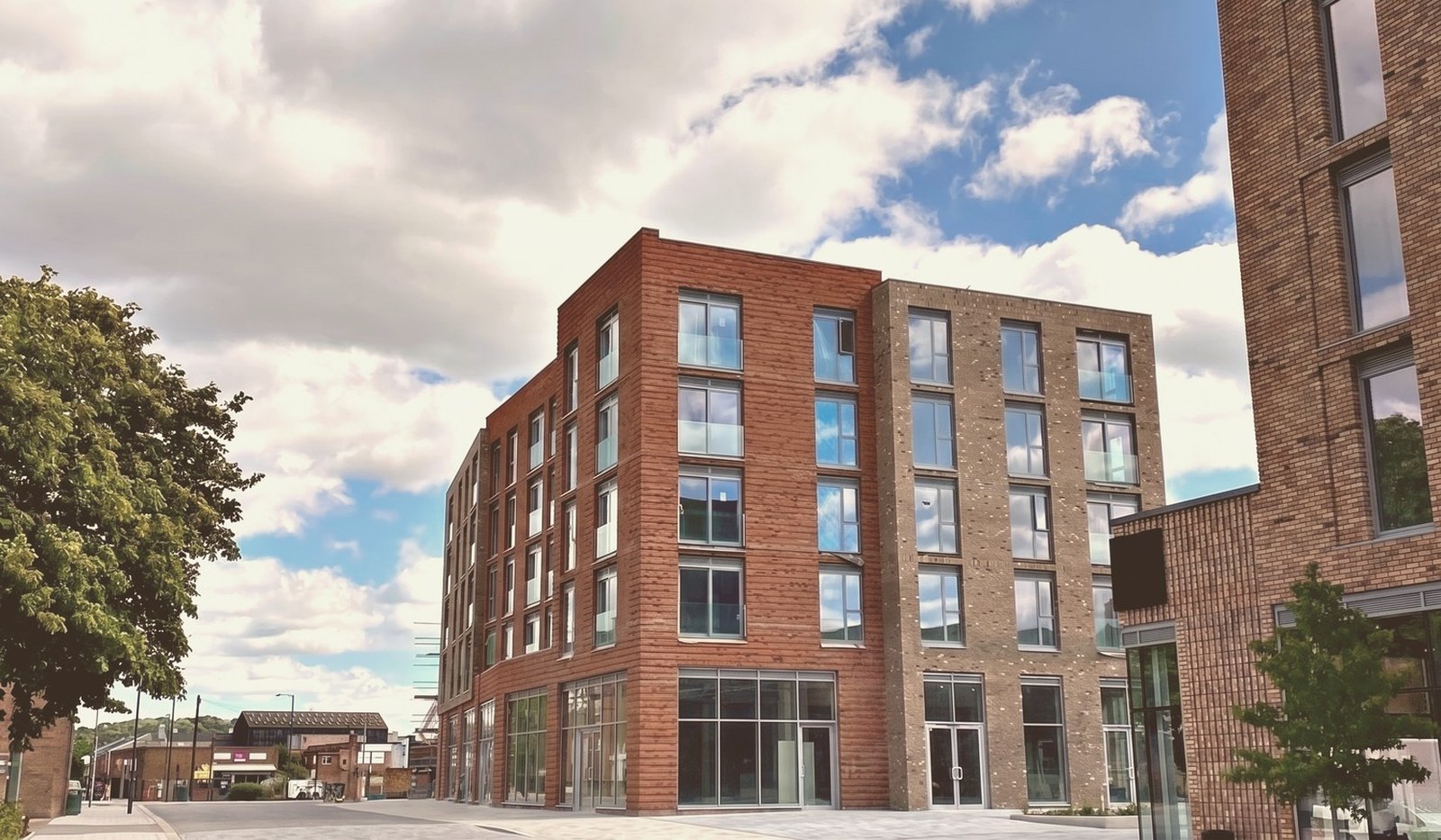 Photograph of the completed five-storey apartment block at Muller Yard, Nottingham: red and buff facing brick with recessed banding, full-height glazed shopfronts at ground level and a chamfered corner turning from Meadow Lane