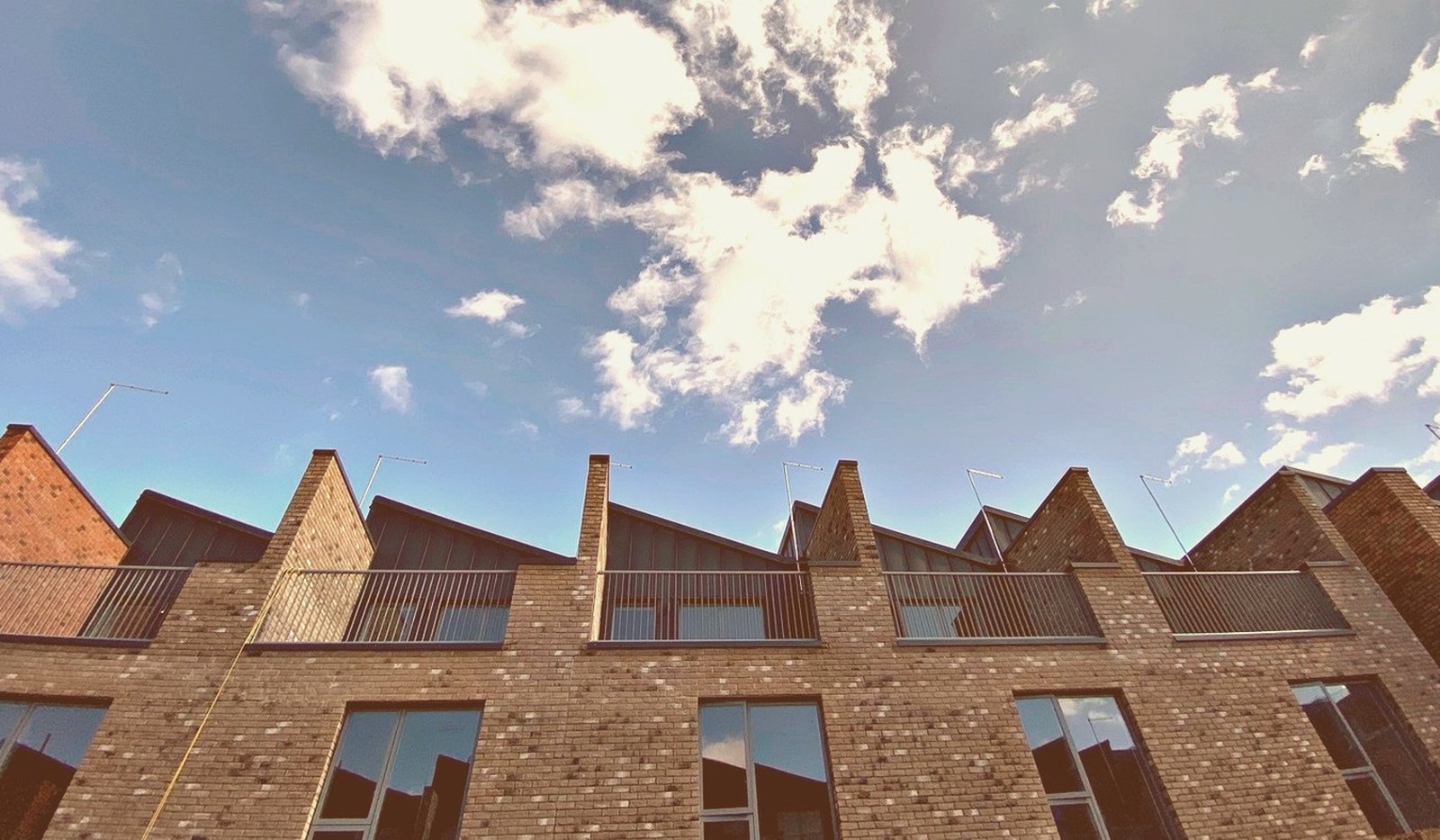 Photograph looking up at the completed townhouse terrace at Muller Yard, Nottingham: a run of asymmetric saw-tooth gables in brick and standing-seam metal against open sky, with vertical screens across the recessed balconies below