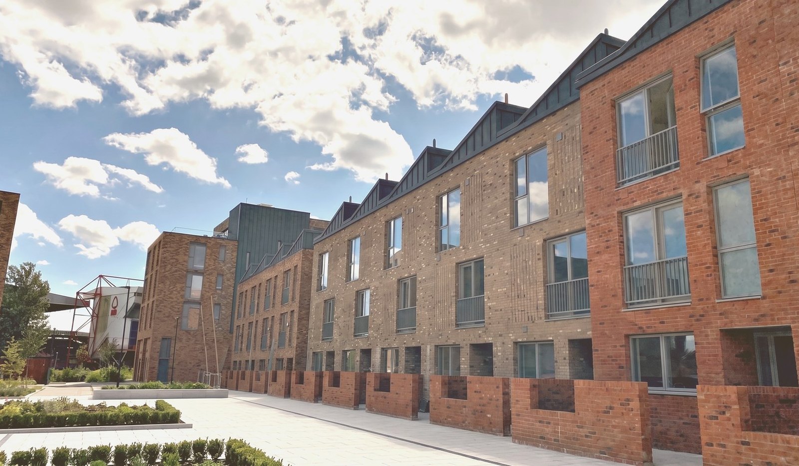 Photograph of the completed townhouse terrace at Muller Yard, Nottingham: brick fronts with recessed balconies and saw-tooth roofs running along one side of a paved parking court, with planted beds and the apartment blocks beyond