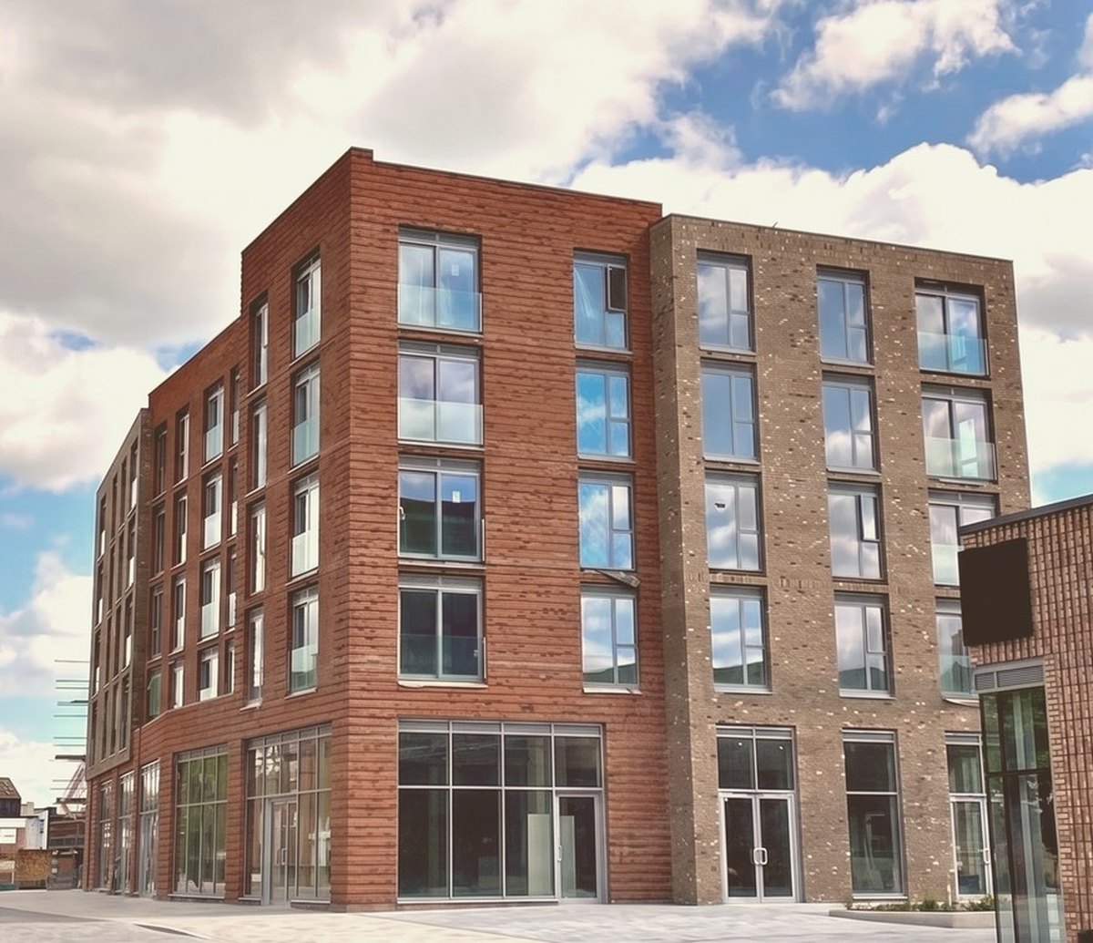 Photograph of the completed Meadow Lane frontage at Muller Yard: the ground floor raised above street level behind a brick-faced wall, with glazed shopfronts set back and the brick apartment storeys rising above