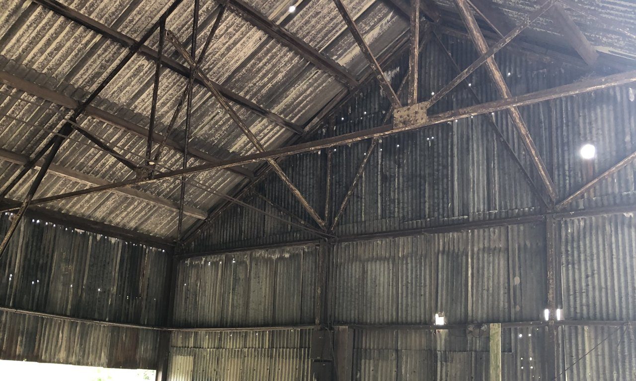 Photograph of the interior of the redundant agricultural barn at Green Gate Barns before conversion: a steel portal frame with light lattice trusses spanning the full width, weathered corrugated metal sheet to the walls and roof, daylight coming through gaps at the eaves, and farm plant standing on the hardstanding below