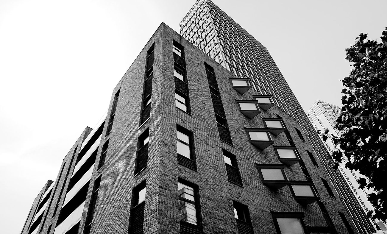 Photograph of the completed building at Tennant Street, Birmingham: the brick corner rising against a bright sky, with angled oriel bay windows stacked up the flank, recessed brick panels between them, and a taller glazed residential tower behind