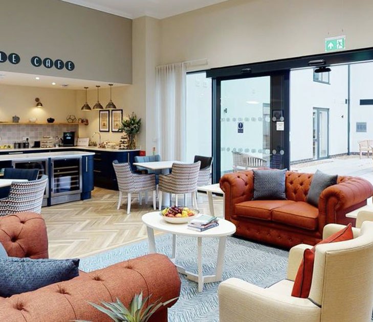Photograph of the Maypole cafe in the completed Brackley Care Home: a servery with pendant lights and stools along a counter, armchairs and a buttoned leather sofa around low tables, and full-height glazed doors opening to the courtyard beyond