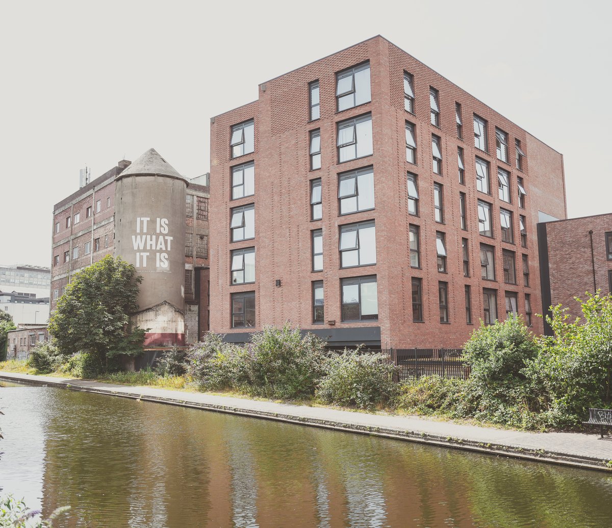 Photograph of the completed Cliveland Street building from the canal towpath: red brick elevations with tall windows rising directly above the water, the retained concrete shot tower alongside and reflections along the canal