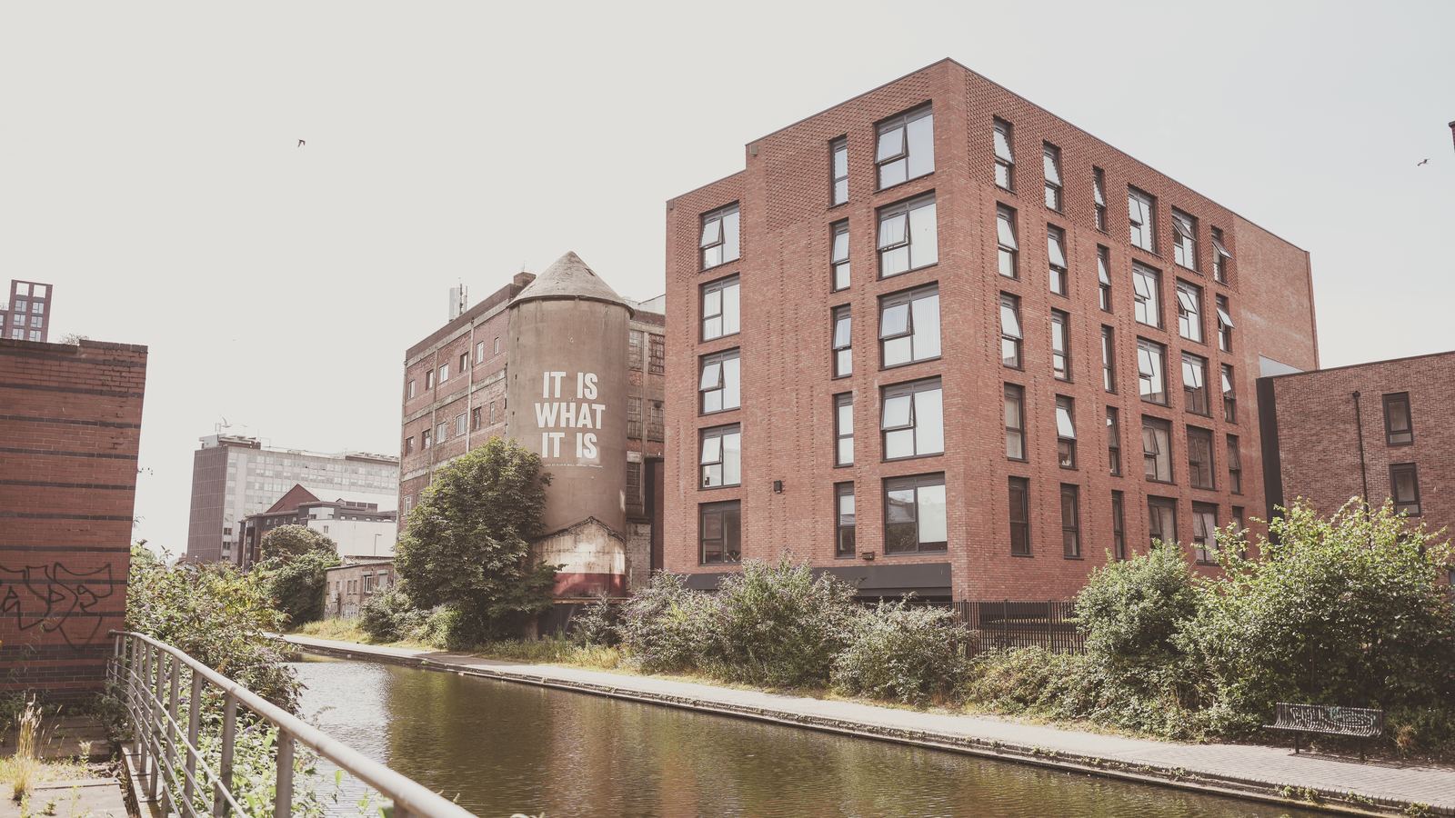 Photograph of the completed apartment building at Cliveland Street, Birmingham: a six-storey red brick block standing along the Birmingham and Fazeley Canal, with tall regular windows, a set-back top storey, the locally listed concrete shot tower beside it and the towpath running along the near bank