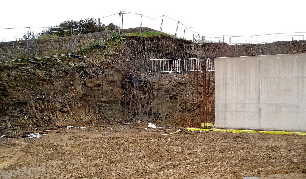 Photograph of the cut cliff face at Gara Rock beside a new in-situ concrete retaining wall holding back the excavated ground, construction fencing along the top of the cut