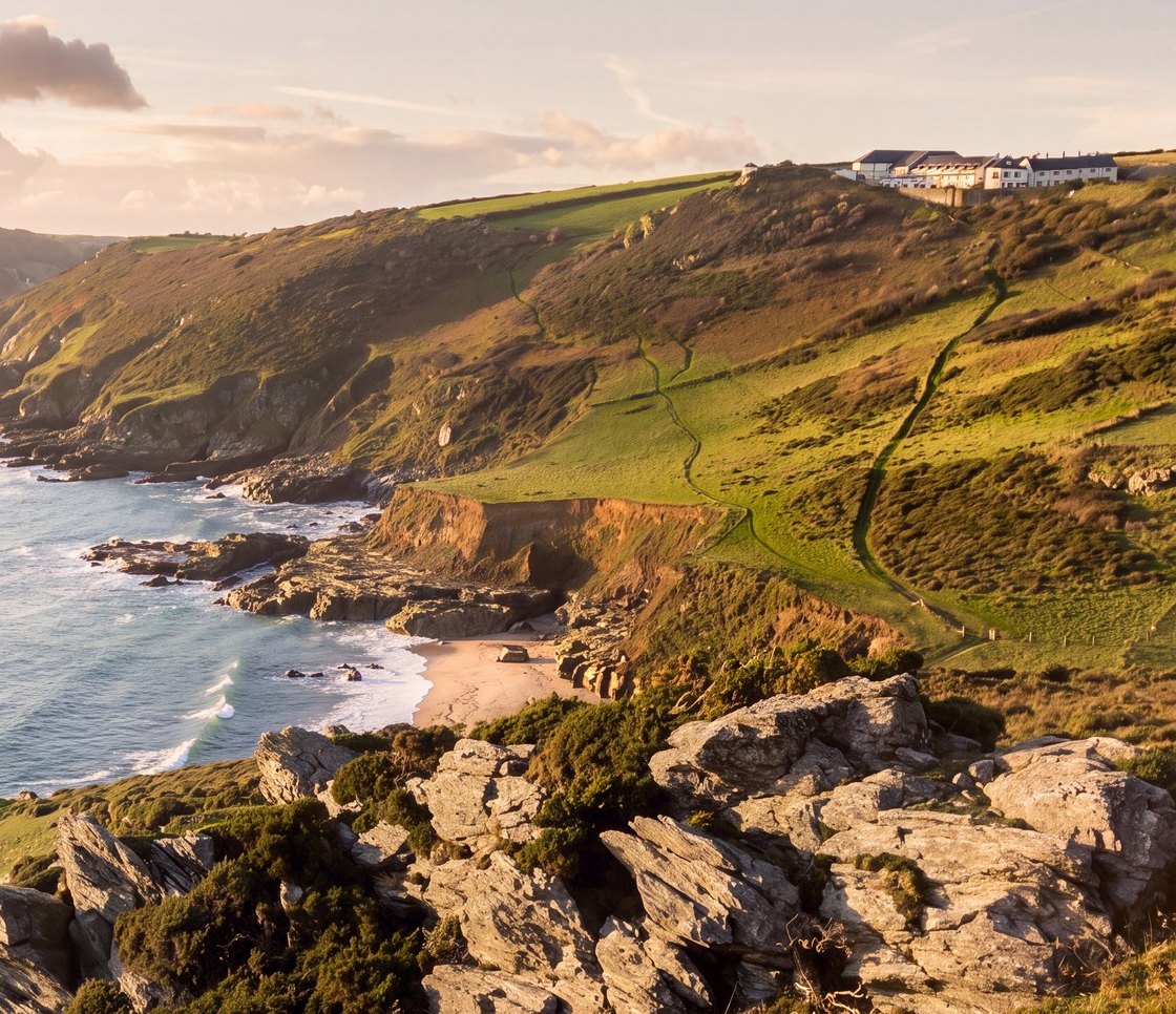 Rickett Architects visualisation of the Gara Rock setting: the cliff-top scheme distant on the headland above the South Devon coast at golden hour, with a rocky foreshore and a sandy beach cove below