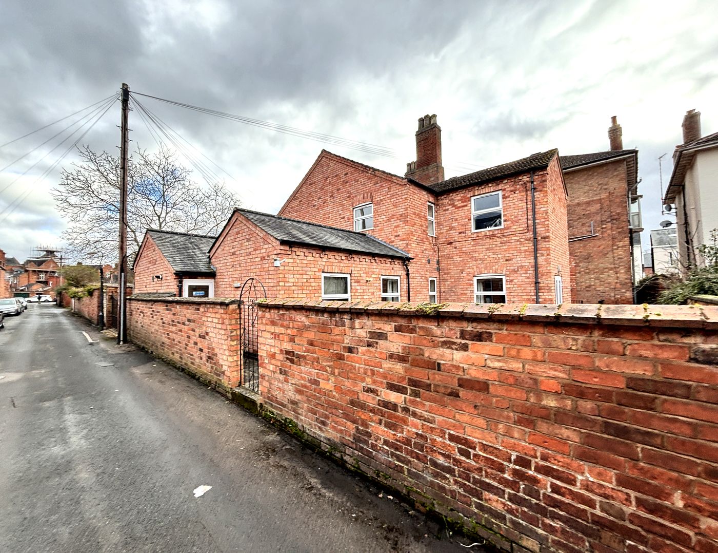 Photograph looking along Gordon Street at the rear of 59 Russell Terrace, Leamington Spa: a red brick boundary wall running down the back lane with a narrow wrought-iron pedestrian gate set into it, the slate-roofed single-storey rear additions behind, and the two-storey brick rear of the villa with its chimney stack