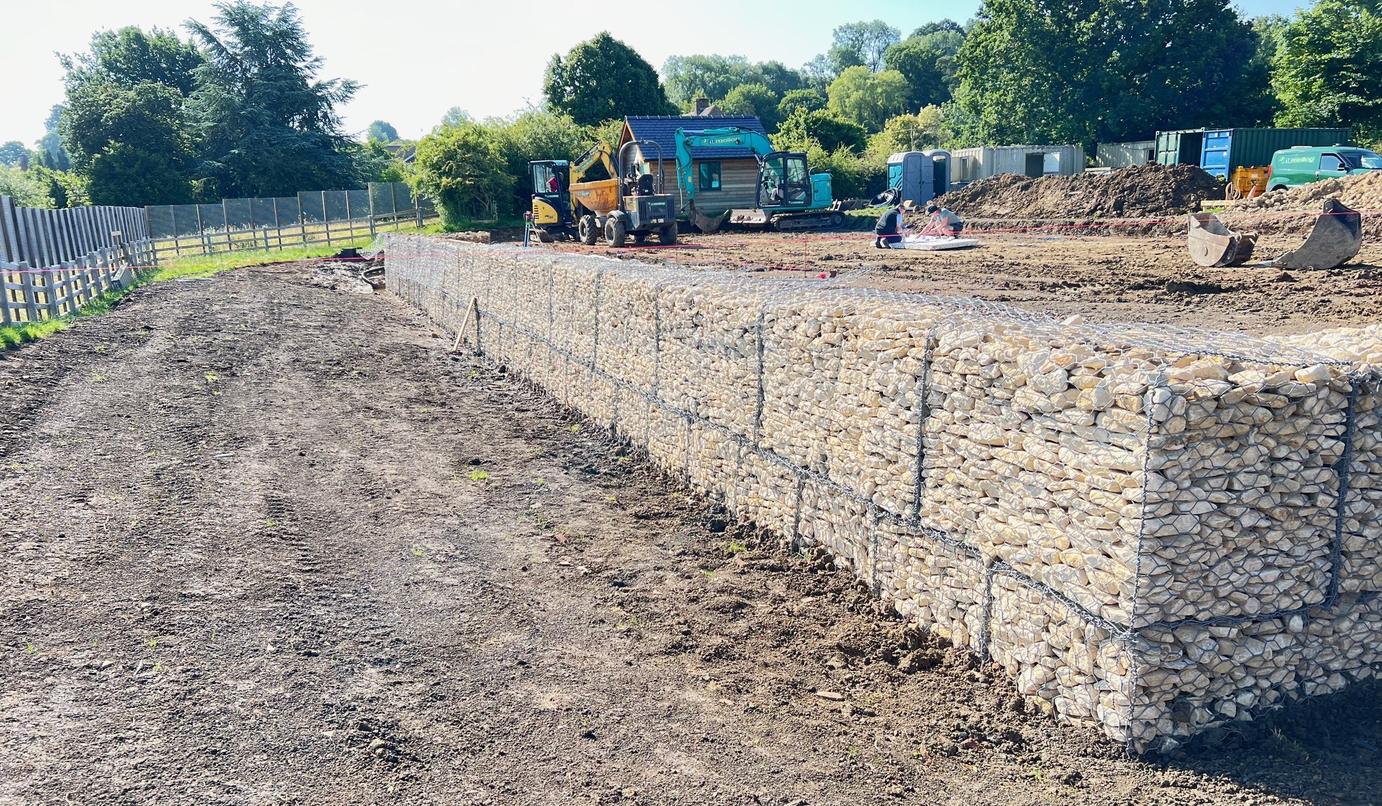 A long run of stone-filled gabion cages retaining the cut across the site at Hollow Meadow Farmhouse during construction, with plant working on the levelled platform beyond