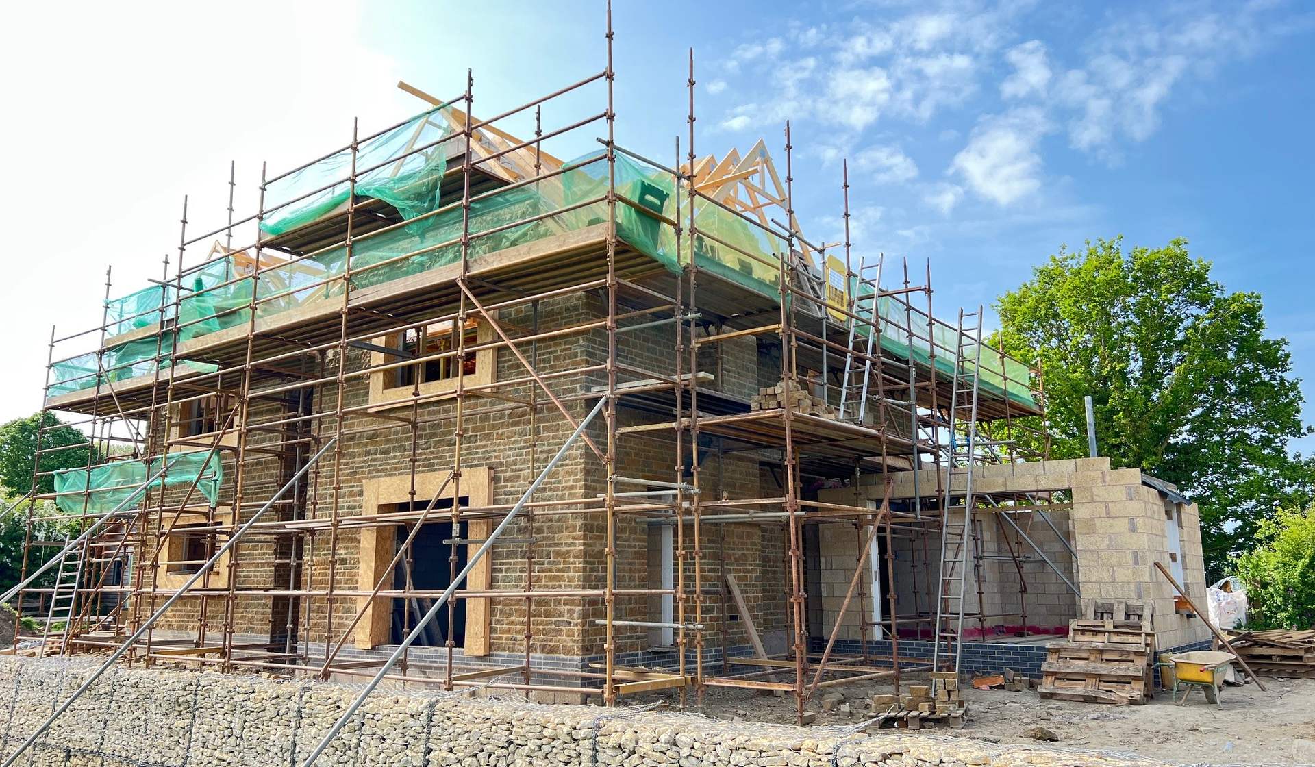 Hollow Meadow Farmhouse under scaffolding during construction, its walls rising in coursed natural ironstone with the roof structure going on above and stone lintels set over the openings