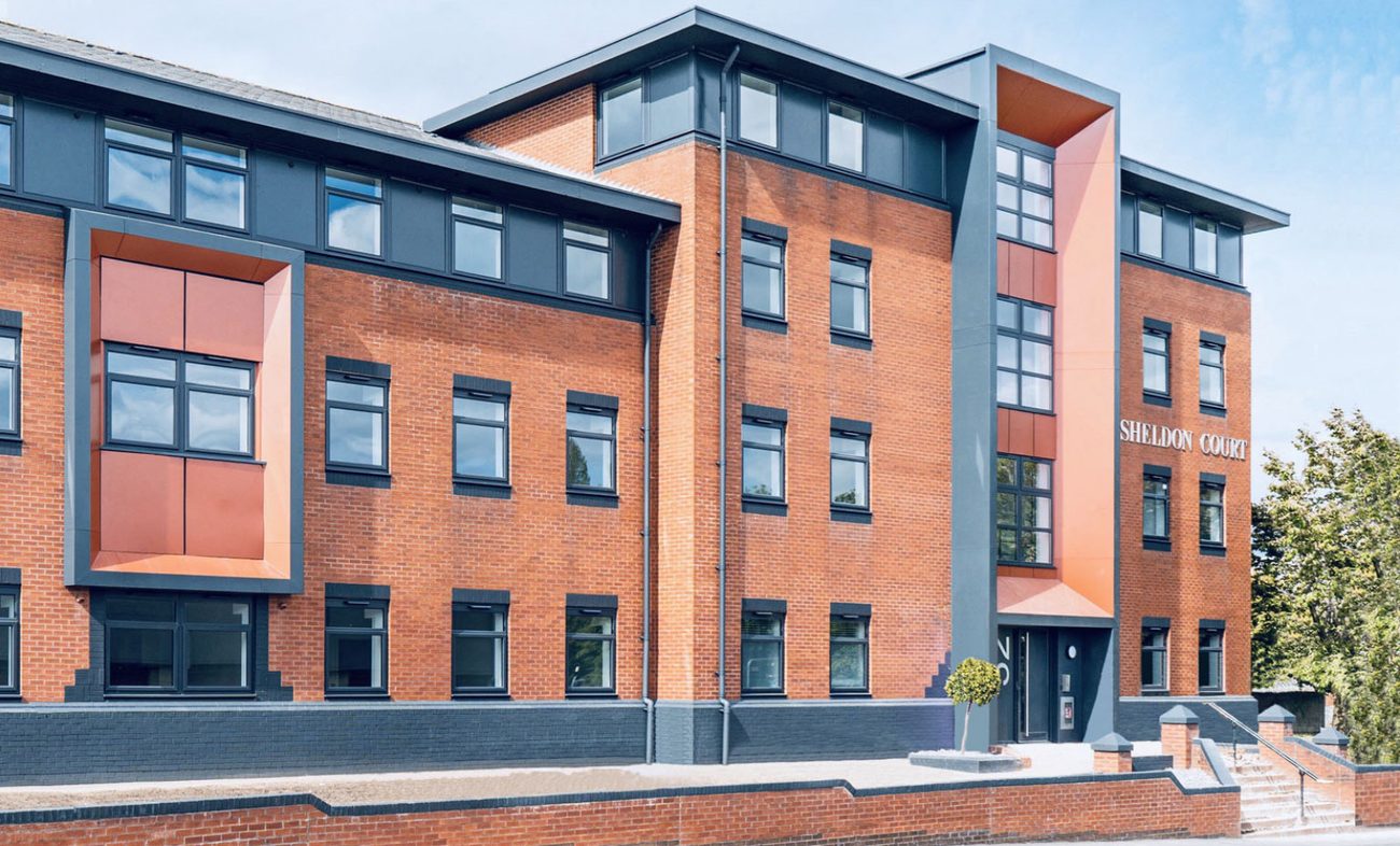 Photograph of the completed Sheldon Court from Wagon Lane: three storeys of red brick with anthracite window surrounds and a dark painted plinth, an orange clad panel set into the elevation, the projecting entrance feature carrying the Sheldon Court lettering, and a young tree and steps at the entrance